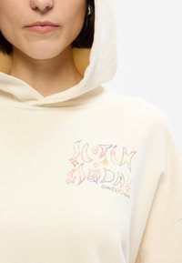 Κρεμ καπιτονέ φούτερ με απαλή υφή, με πολύχρωμο γραφικό στην αριστερή πλευρά που περιλαμβάνει κείμενο και διακοσμητικά στοιχεία.