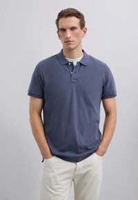 Polo shirt bleu marine à manches courtes, avec patte de boutonnage à deux boutons et tissu côtelé texturé. Associé à un pantalon couleur crème, en position debout.