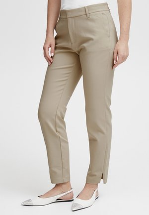Personne portant un pantalon beige slim jusqu’aux chevilles avec couture devant, fente latérale à l’ourlet, et des ballerines blanches pointues à bouts cloutés, debout sur fond blanc.