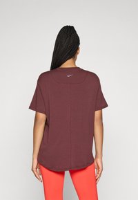 Donna con lunghe trecce che indossa una t-shirt Nike larga color bordeaux e leggins arancioni brillanti, vista di spalle su uno sfondo neutro.