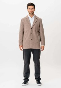 Mand står iført en beige blazer, hvid skjorte, mørkegrå bukser og sorte sneakers foran en ensfarvet hvid baggrund.
