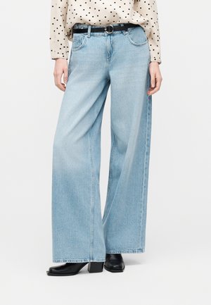 Φαρδιά τζιν σε ανοιχτό γαλάζιο χρώμα με πέντε τσέπες, φορεμένα με μαύρη ζώνη, μαύρες μπότες με τακούνι και ένα κρεμ πουκάμισο με πουά, που είναι βιδωμένο μέσα.