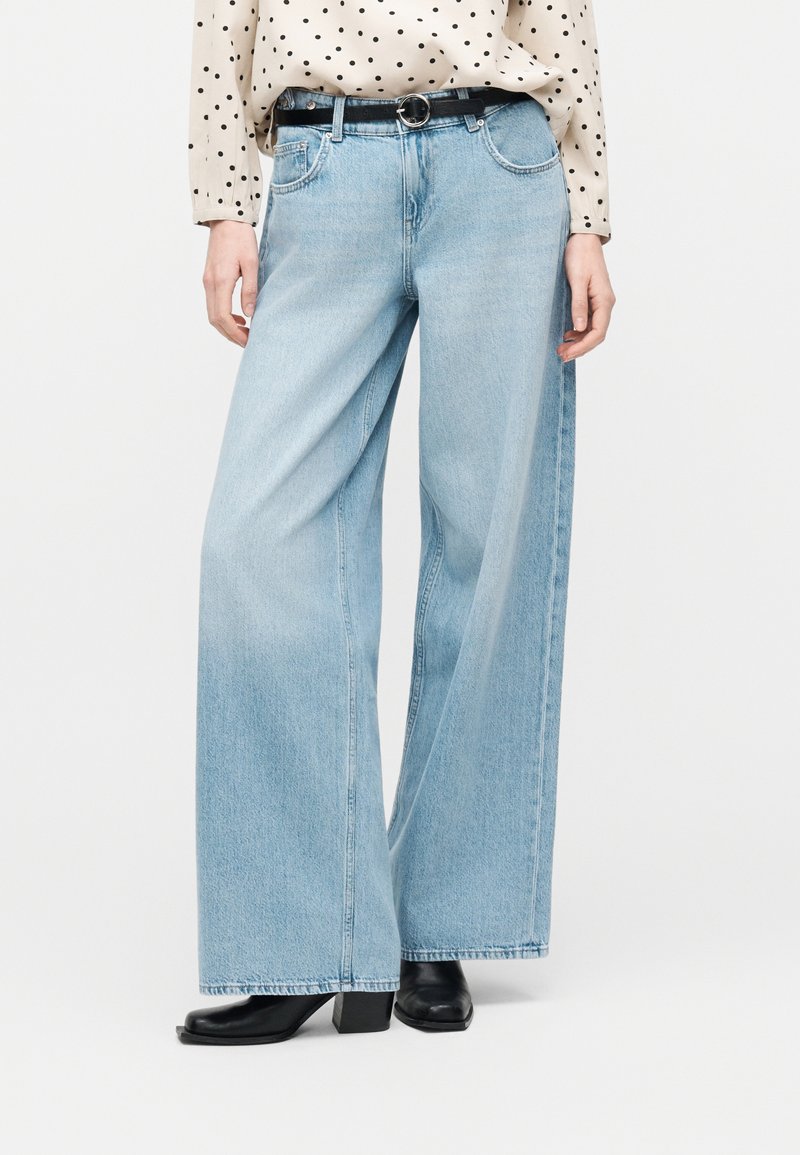 Φαρδιά τζιν σε ανοιχτό γαλάζιο χρώμα με πέντε τσέπες, φορεμένα με μαύρη ζώνη, μαύρες μπότες με τακούνι και ένα κρεμ πουκάμισο με πουά, που είναι βιδωμένο μέσα.