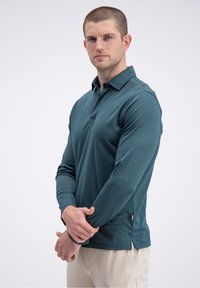Tamsiai žalsvai mėlynas ilgomis rankovėmis polo marškinėlis su apykakle, mygtukų skylių juostele ir šoninėmis įpjovomis. Minkšta medžiaga su subtiliu blizgesiu ir mažu logotipu krūtinės srityje.