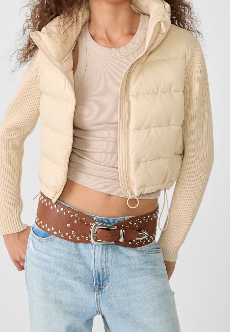 Femme portant une doudoune courte beige, manches longues côtelées, débardeur beige, ceinture en cuir marron cloutée, et jean bleu clair.