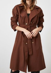 Hnědý trench coat s páskem v pase, dvojitými knoflíky, oversized klopami a ohrnutými rukávy, vyrobený z hladké tkaniny.