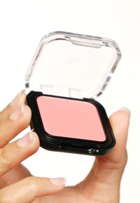 Poudre à blush compacte avec un couvercle transparent, une base noire, présentant un godet de blush rose doux. La compacte est tenue entre les doigts, mettant en valeur sa forme rectangulaire.