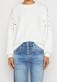 Bílá mikina se střiženým květinovým vzorem na rukávech, s kulatým výstřihem a uvolněným střihem. Kombinováno s modrými džíny s vysokým pasem.