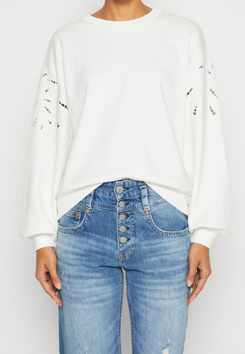 Bílá mikina se střiženým květinovým vzorem na rukávech, s kulatým výstřihem a uvolněným střihem. Kombinováno s modrými džíny s vysokým pasem.