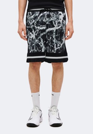 Homme portant un short de basket noir et blanc à motif, des chaussettes blanches Nike et des chaussures de sport blanches Nike, debout devant un fond uni.