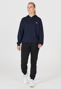 Marinblå huvtröja med en liten logotyp, ihop med svarta avsmalnande byxor och vita sneakers. Tyget ser slätt ut med långa ärmar.