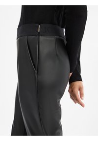 Ingrandimento di una persona che indossa pantaloni in pelle sintetica nera con vita alta e logo del marchio visibile, abbinati a un top nero a maniche lunghe.