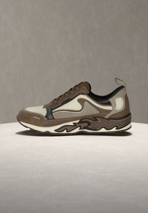 Bruine en beige sportsneaker met meshpanelen, veterontwerp en patroon op de zool, geplaatst op een lichte ronde ondergrond tegen een neutrale achtergrond.