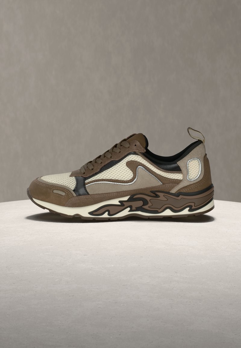 Καφέ και μπεζ αθλητικό παπούτσι με διχτυωτές επιφάνειες, δεσίματα με κορδόνια και σόλα με σχέδιο, τοποθετημένο σε ελαφρώς στρογγυλή επιφάνεια πάνω σε ουδέτερο φόντο.