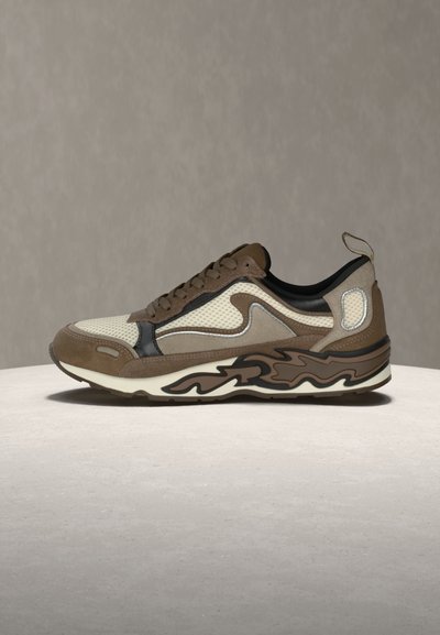 Καφέ και μπεζ αθλητικό παπούτσι με διχτυωτές επιφάνειες, δεσίματα με κορδόνια και σόλα με σχέδιο, τοποθετημένο σε ελαφρώς στρογγυλή επιφάνεια πάνω σε ουδέτερο φόντο.