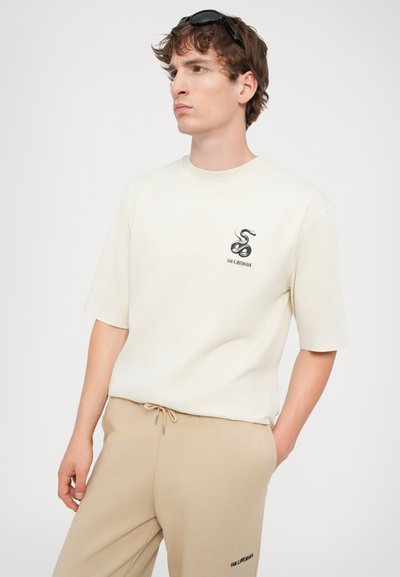 T-shirt crème à manches courtes avec un graphique de serpent noir et un logo. Associé à un pantalon beige à cordon de serrage portant le nom de la marque imprimé.