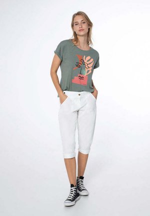 Camiseta corta de manga verde con un diseño gráfico en coral, combinada con pantalones cortos blancos y zapatillas negras de caña alta. Atuendo simple y casual.