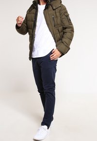 Olivgrön puffjacka med dragkedja framtill och huva, bärs över en vit t-shirt och marinblå byxor, ihop med vita sneakers.