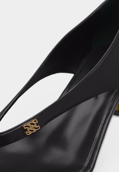 Μαύρο ψηλοτάκουνο παπούτσι με μυτερή μύτη, με χρυσό διακοσμητικό λογότυπο στο λουράκι στο πάνω μέρος.