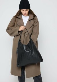 Schwarze Leder-Schultertasche mit einem gesteppte Muster, verjüngter Form und zwei Schulterriemen. Wird getragen von einer Person in brauner Jacke und Mütze.