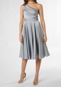 Žena v stříbrných saténových midi šatech na jedno rameno s plisovaným tělem a rozšířenou sukní, doplněna stříbrnými sandály na vysokém podpatku.