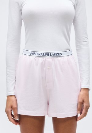Femme portant un short rose clair avec une ceinture marquée Polo Ralph Lauren et un haut blanc ajusté à manches longues, debout les mains détendues.