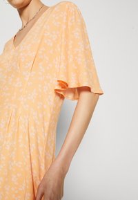 Robe florale orange avec un décolleté en V, de courtes manches évasées, un tissu doux et des plis à l'avant. Présente de petits motifs de fleurs blanches sur l'ensemble.