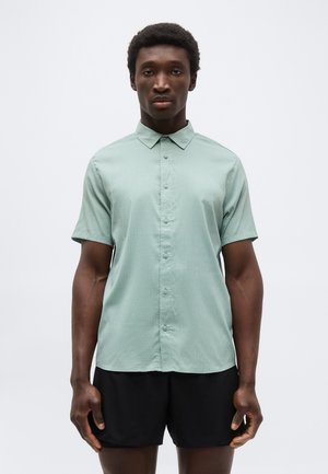 Άνδρας με ανοιχτοπράσινο πουκάμισο με κοντά μανίκια και κουμπιά και μαύρο σορτς, στέκεται μπροστά σε απλό λευκό φόντο, κοιτάζοντας μπροστά.