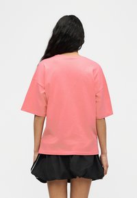 Άτομο με μακριά σκούρα μαλλιά, φοράει φαρδιά ροζ κοντομάνικη μπλούζα και μαύρο σορτς, στέκεται γυρισμένο με την πλάτη σε ένα απλό φόντο.