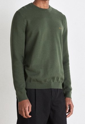 Homme portant un pull vert olive à manches longues avec col rond et logo "BOSS" sur la poitrine, et un pantalon noir, debout devant un fond uni.
