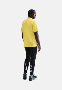 Polo shirt jaune à manches courtes avec des accents verts, associé à un pantalon noir avec un logo Nike blanc et le texte "AIR", et des chaussures de sport.