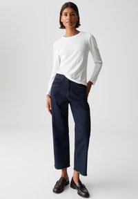 Donna con capelli corti scuri che indossa una camicia bianca a maniche lunghe, jeans blu scuro tagliati in fondo e mocassini marroni scuri, in piedi davanti a uno sfondo grigio.