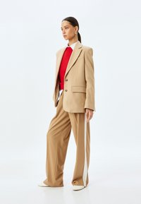 Beige op maat gemaakte blazer met twee knopen, gecombineerd met wijde beige broeken met een witte streep. Rode gebreide trui eronder, gouden oorbellen.