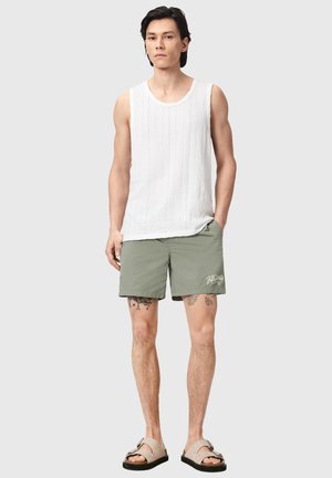 Man staat en draagt een wit mouwloos geribd shirt, olijfgroene shorts met logo, beige sandalen, en zichtbare tatoeages op de dijen.