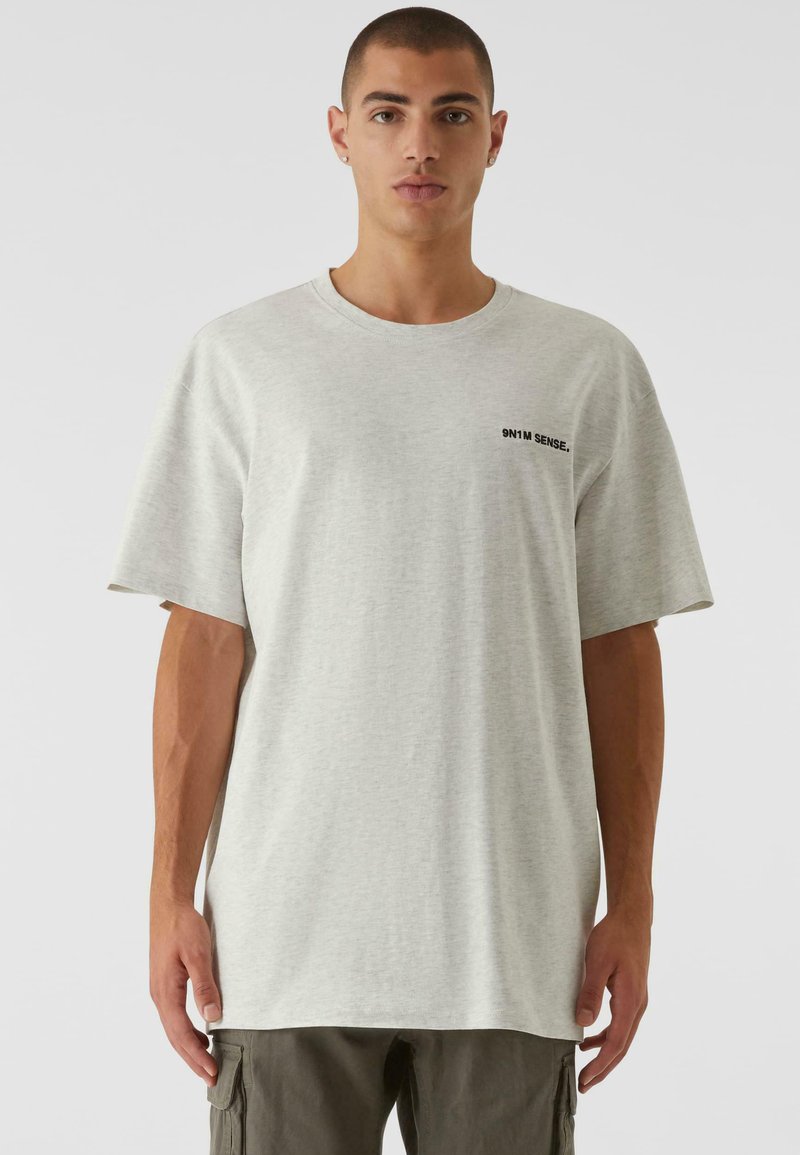 Lys grå oversized t-shirt lavet af bomuldsblanding, med rund halsudskæring og korte ærmer, og lille sort tekst på venstre bryst.