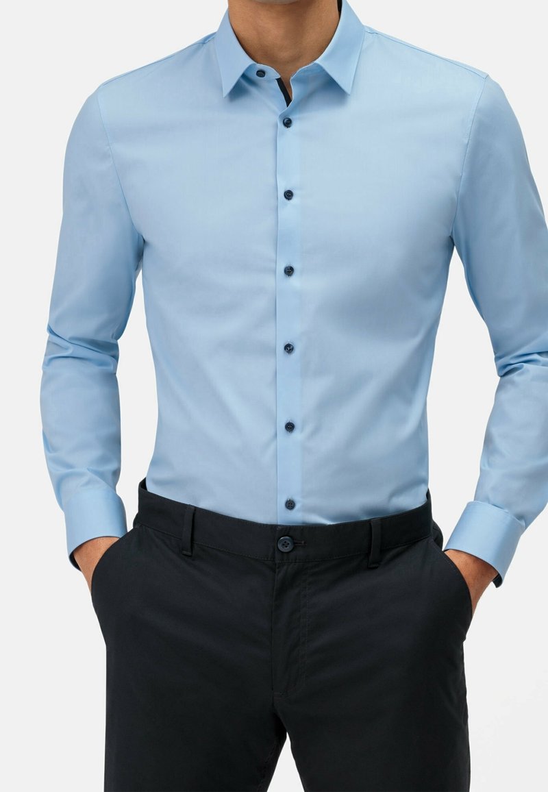 Chemise bleu clair à manches longues avec une coupe semi-ajustée et des boutons foncés, associée à un pantalon foncé.