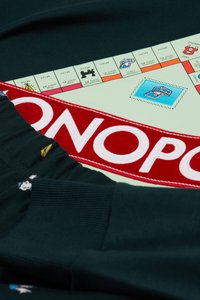 Tejido negro con un logotipo de Monopoly en bordado rojo y blanco, con detalles coloridos de personajes y un borde decorado con un patrón de un tablero de Monopoly.