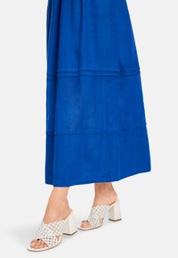 Weiße, offene Mules mit einem geflochtenen Muster und einem klobigen Absatz. Getragen mit einem fließenden, leuchtend blauen Maxikleid mit horizontalen Nähten.