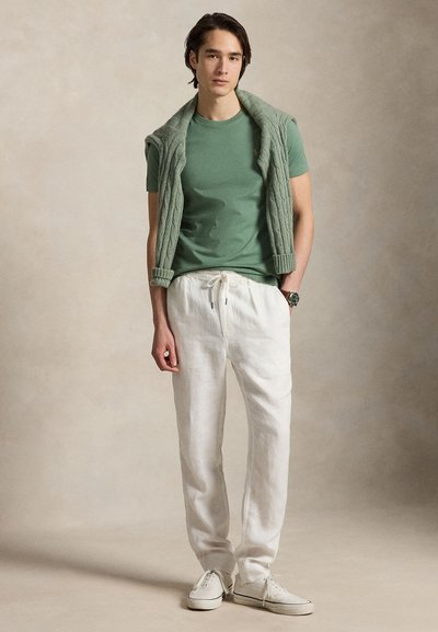 Mlad moški nosi žajbljevo zeleno majico, bele hlače z vrvico, bele superge in zelen pulover z pletenim vzorcem, prevržen čez ramena.