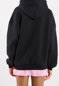 Černá oversize mikina s klokaní kapsou a kapucí na tkaničku. Měkká látka, žebrovaný lem a manžety. Růžové šaty vykukující spodem.