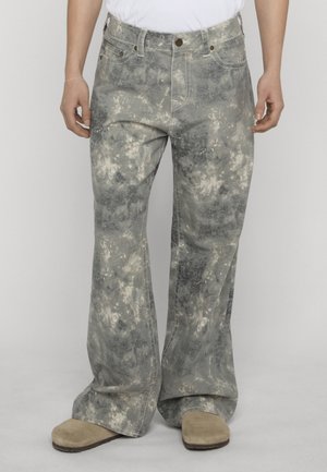 Uomo che indossa jeans a gamba larga grigio e bianco tie-dye con scarpe beige e una camicia bianca, in piedi contro uno sfondo semplice.