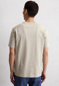 Beige T-Shirt mit kurzen Ärmeln aus weicher Baumwolle, mit Rundhalsausschnitt und lockerer Passform, getragen mit blauen Denim-Hosen, von hinten betrachtet.