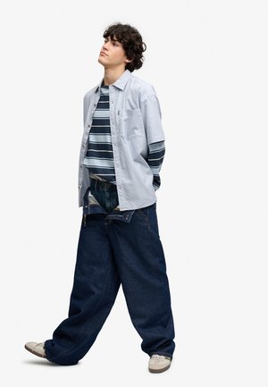 Ung person står med hænderne bag ryggen iført oversized mørkeblå jeans, stribet skjorte, lys åben skjorte med knapper og beige sneakers.