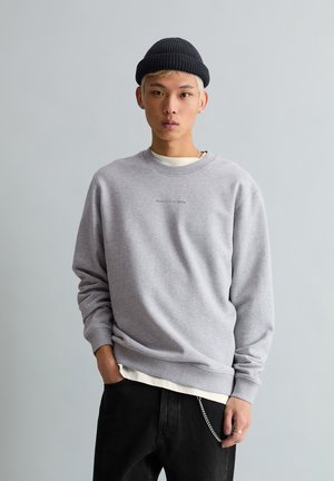 Jeune personne aux cheveux blonds courts portant un bonnet noir, un sweat-shirt gris, un sous-pull blanc, un pantalon noir avec une chaîne, debout devant un fond gris.