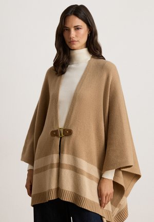 Femme aux cheveux foncés portant un poncho beige en maille avec une boucle en cuir au-dessus d’un pull à col roulé blanc et d’un pantalon foncé.