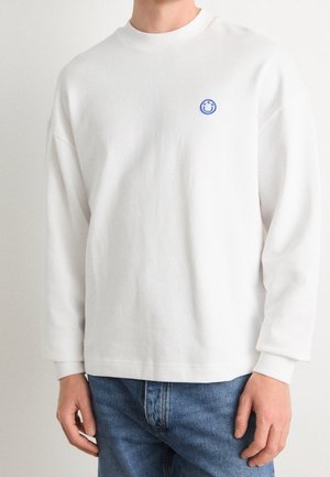 Biała koszula z długim rękawem z małym niebieskim logo uśmiechniętej buźki na lewej piersi, noszona przez osobę w niebieskich dżinsach na jednolitym tle.