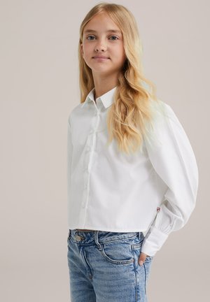 Blondes Mädchen mit langen, gewellten Haaren, das eine weiße Bluse mit Puffärmeln und blaue Jeans trägt und mit der Hand in der Tasche steht.
