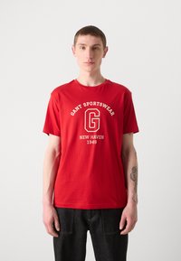 Rød bomulds T-shirt med rund halsudskæring og korte ærmer. Trykt hvid branding med et logo, placering og årstal foran.