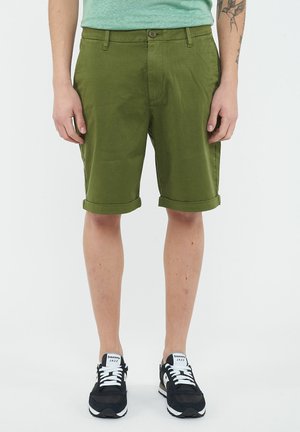 Adulto che indossa pantaloncini verde oliva arrotolati, scarpe da ginnastica Saucony Jazz nere e bianche, in piedi con le mani rilassate lungo i fianchi.