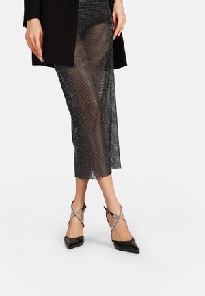 Décolleté nere con punta a punta caratterizzate da una finitura lucida e da cinturini incrociati argentati. Il modello indossa una gonna con sovrapposizione trasparente a pois.